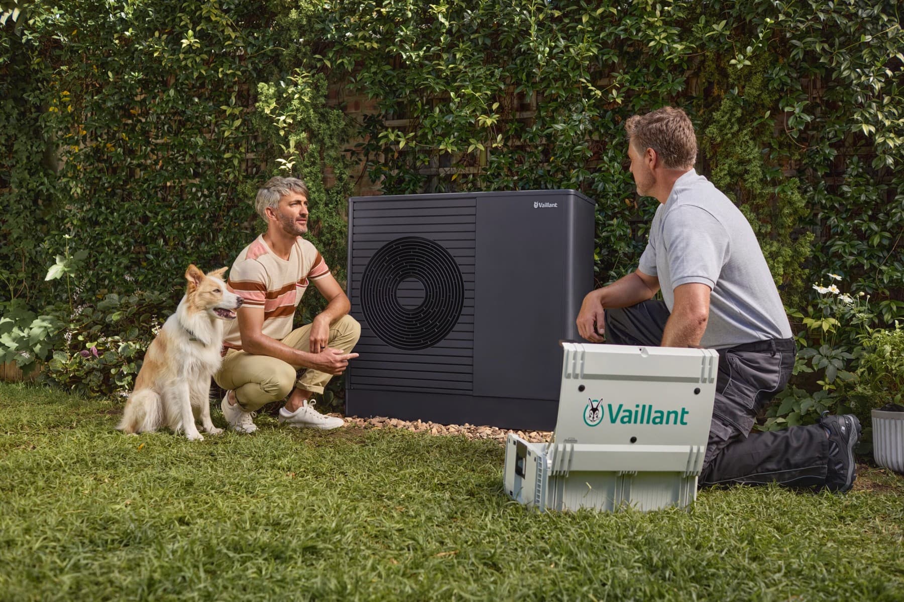 Een installateur zit gehurkt voor een warmtepomp en legt iets uit aan een man.